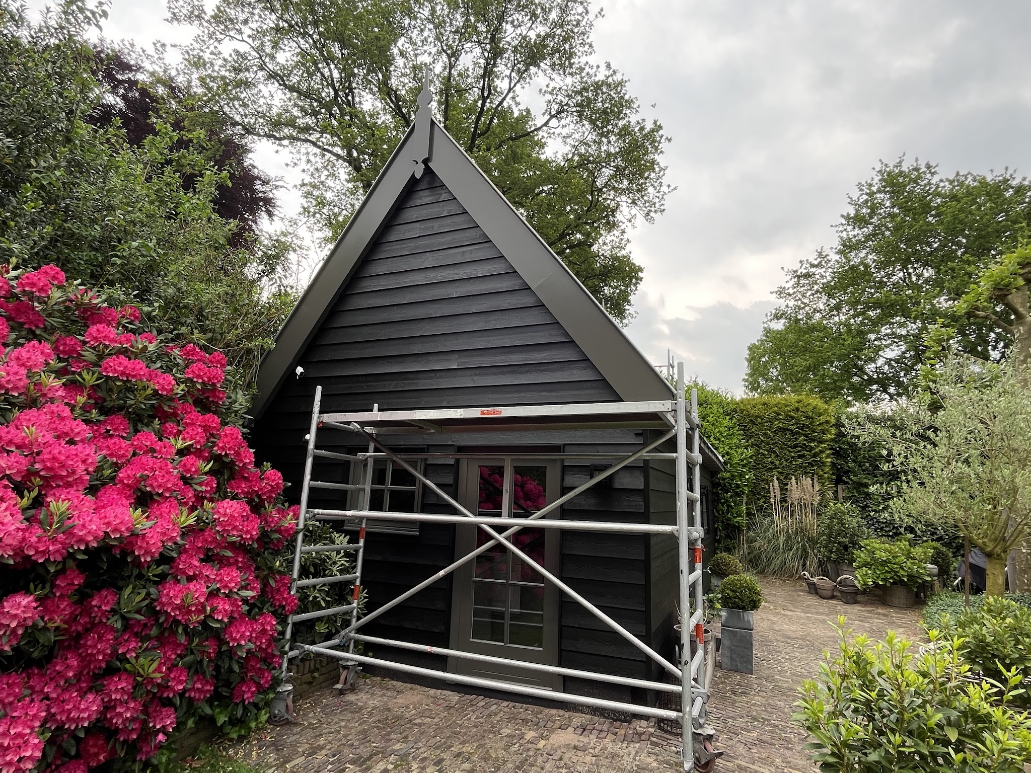 Zwart houten tuinhuis met puntgevel, rolsteiger ervoor en bloeiende rododendron ernaast